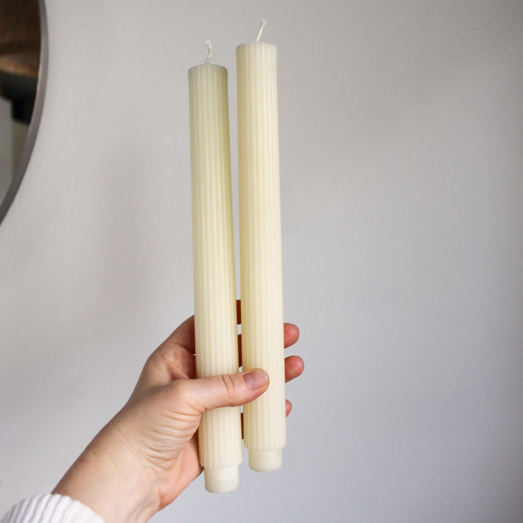 A pair of beeswax taper candles held up by a light-skinned hand against a white wall.