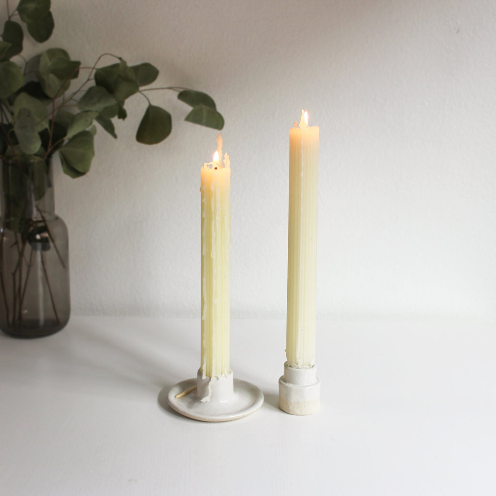 A pair of taper candle holders made of white clay with white glaze holding lit beeswax taper candles on a white table.
