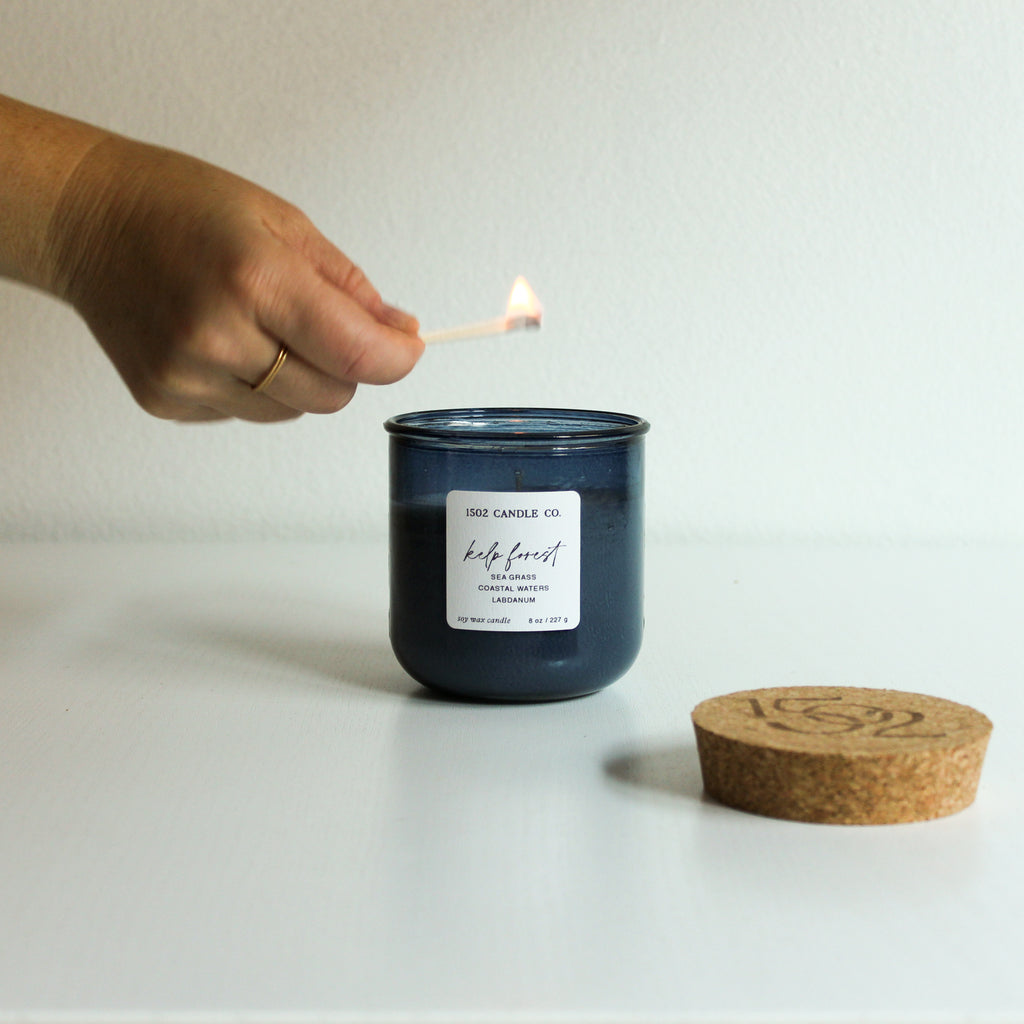 Hand lighting a moody ocean blue candle labeled 'Kelp Forest' with a natural 
cork topper on a white surface.