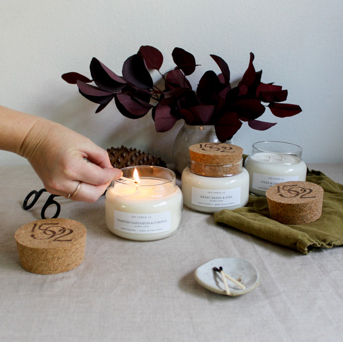 a light skinned hand lighting a candle next to a Sweet Berry and Pine apothecary jar soy wax candle on a table