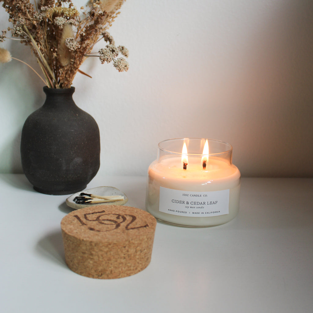 a lit Cider and Cedar Leaf apothecary jar soy wax candle on a white table