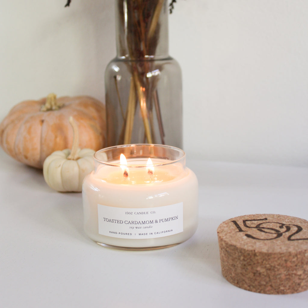 a lit Toasted Cardamom and Pumpkin apothecary jar soy wax candle on a white table next to pumpkins