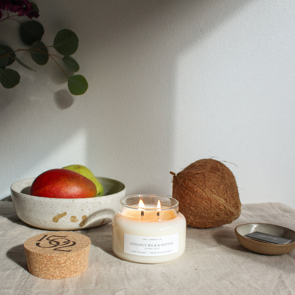 Coconut Milk and Nectar apothecary jar soy wax candle lit on a table in front of a coconut and a bowl filled with fruit.
