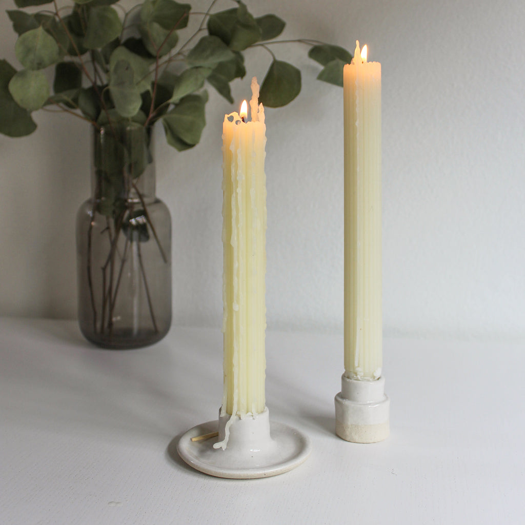 A pair of lit beeswax taper candles on a white table next to a vase with dried eucalyplus.