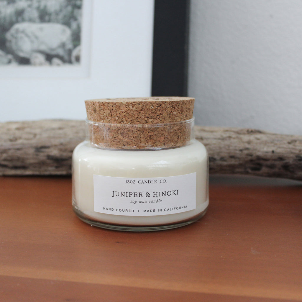 Juniper and Hinoki apothecary jar soy wax candle on a wood shelf in front of a branch resting against black picture frame.