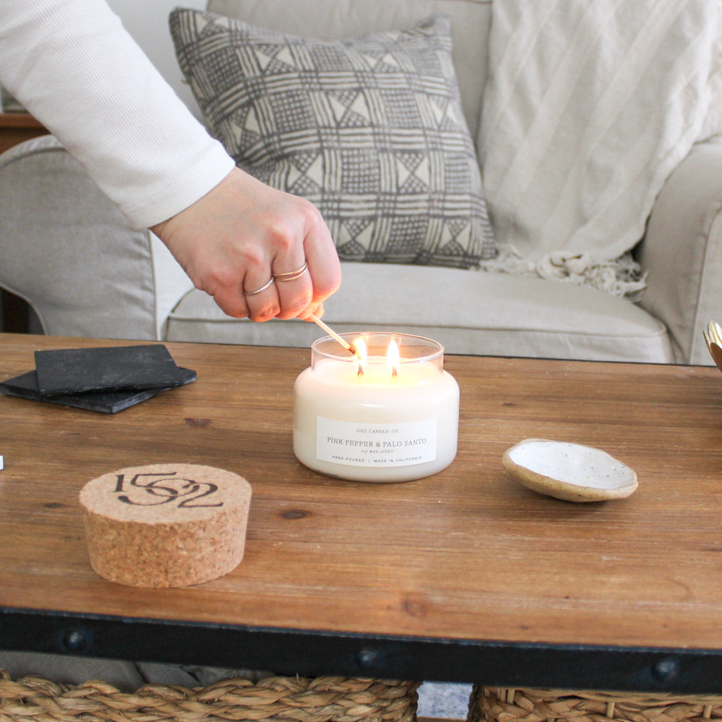 A Pink Pepper and Palo Santo soy wax candle is lit by a light skined had in a California Modern styled livingroom.