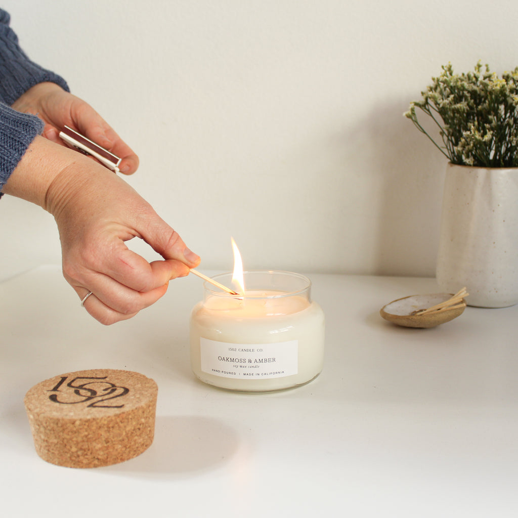 A natural soy wax candle is lit on a white table by a light skined hand.